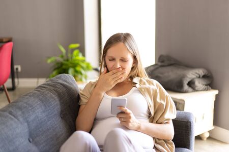 Sleepy pregnant woman checking social media chats on smart phone. Expectant mother sitting on couch,...
