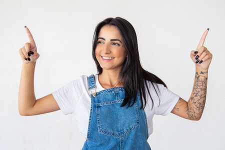 Joyful brunette pointing with fingers up. Studio shot of cheerful young woman wearing denim jumpsuit. Concept of indicationの写真素材