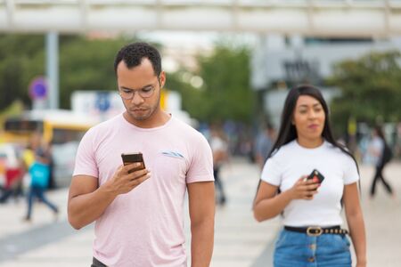 Thoughtful man looking at phone while walking on street. Handsome African American guy strolling...