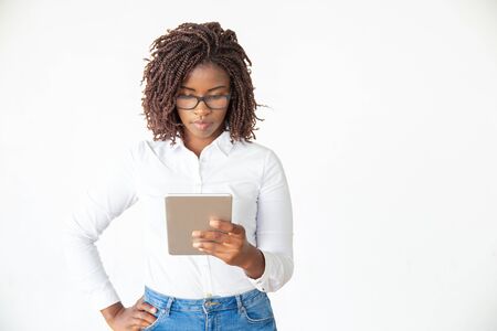 Focused serious expert in glasses using tablet, reading on screen. Young African American female professional standing isolated over white background. Digital communication conceptの写真素材