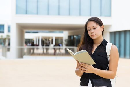 Concentrated young woman reading book. Young female college student with backpack standing on street and reading book. Education conceptの写真素材