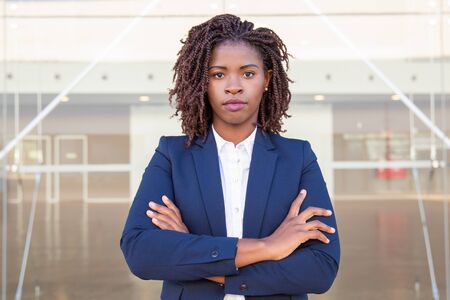 Serious confident business woman posing near office building. Young African American business leader with arms crossed standing outside, looking at camera. Successful businesswoman conceptの写真素材