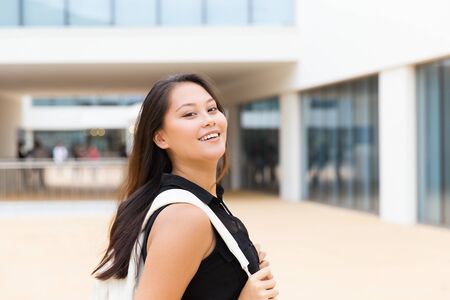 Happy joyful female tourist posing outside. Young mix raced woman in casual standing near city building, adjusting backpack, looking at camera, smiling. Happy young tourist conceptの写真素材