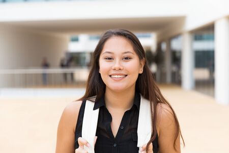 Happy excited female tourist posing outside. Young mix raced woman in casual standing near city building, holding backpack straps, looking at camera, smiling. Young woman portrait conceptの写真素材