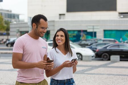 Cheerful friends strolling on street with smartphones. Smiling young people using phones while...