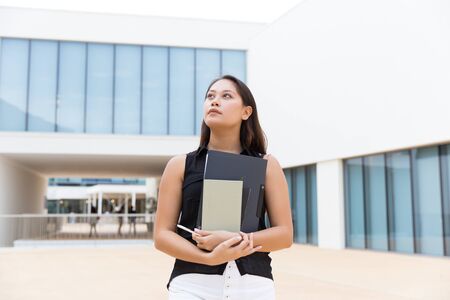 Pensive female intern carrying documents. Young Asian woman walking outside, holding notebook and...
