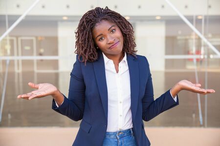 Positive confused female agent posing outside. Young black business woman standing near outdoor glass wall, looking at camera, shrugging. Puzzlement conceptの写真素材
