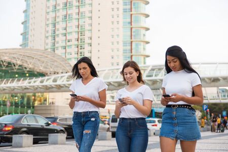 Joyful friends walking on street with smartphones. Smiling young women holding modern phones during stroll. Technology conceptの写真素材