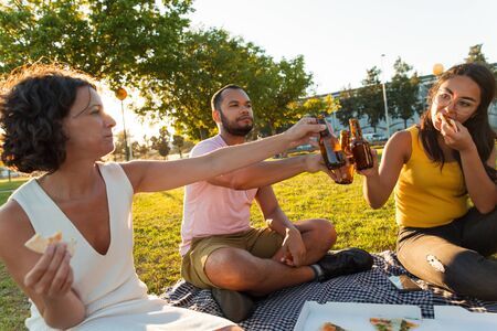 Happy relaxed friends drinking and toasting beer in park. Man and women sitting on grass, eating pizza and clinking bottles. Picnic or party conceptの写真素材