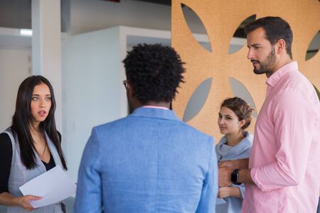 Multiethnic startup team discussing work issues. Young men and women in casual standing together, holding papers, talking. Workgroup communication conceptの写真素材