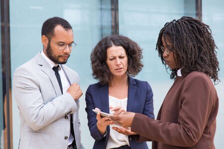 Interracial partners meeting and discussing project outside. Business man and women standing near glass wall, using tablet, watching content together, pointing at screen. Communication conceptの写真素材