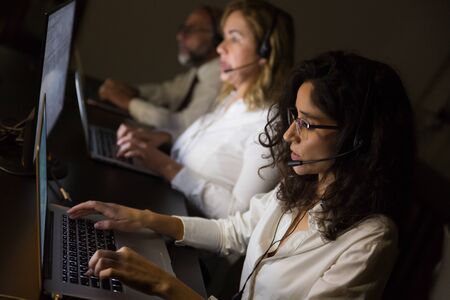 Concentrated call center operators using laptops. High angle view of serious business people in headsets using laptop computers in dark office, selective focus. Technology conceptの写真素材