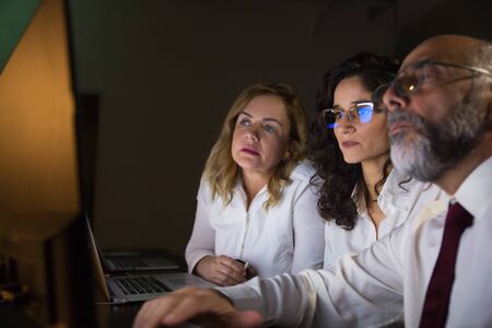 Focused business colleagues working with computer. Side view of focused male and female business people in eyeglasses sitting and using desktop computer in dark office. Working late conceptの写真素材