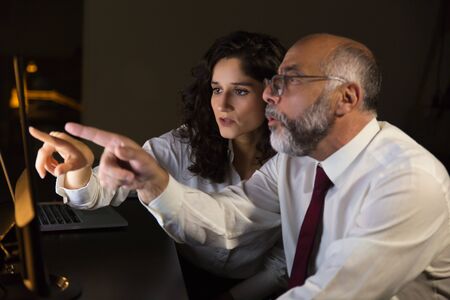 Shocked coworkers pointing at computer monitor. Side view of focused male and female business colleagues looking at computer monitor in dark officeの写真素材