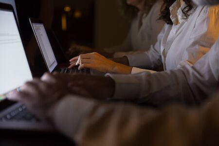 Cropped shot of business people working with laptops. Close-up partial view of professional business colleagues using laptop computers in dark office. Technology conceptの写真素材