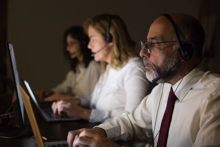 Side view of coworkers in headsets using laptops. Professional focused call center operators in headsets using laptop computers in dark office. Working late conceptの写真素材