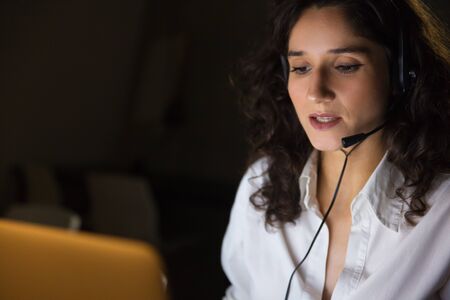Serious young businesswoman in headset. Close-up view of concentrated female call center operator working late in dark office. Client service conceptの写真素材