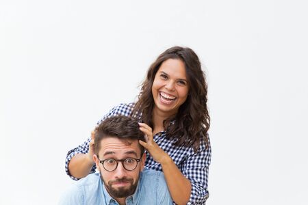Laughing woman setting hair of her scared boyfriend for fun. Young woman in casual and man in glasses standing isolated over white background. Couple having fun conceptの写真素材