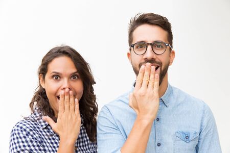 Surprised happy couple staring at camera and covering open mouths with hands. Young woman in casual and man in glasses standing isolated over white background. Great news conceptの写真素材