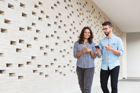 Couple of friends with smartphones showing content on screens to each other. Young man and woman...
