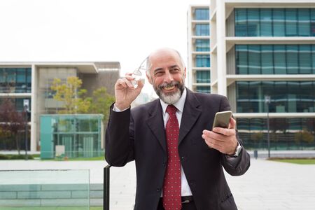 Cheerful businessman holding smartphone and eyeglasses. Happy bearded mature businessman holding mobile phone and spectacles on street. Business success conceptの写真素材