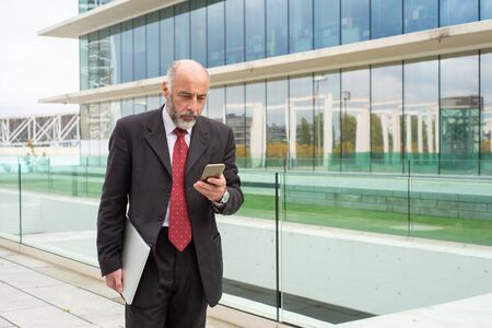 Focused grey haired company owner using smartphone on his way to office. Elderly man in formal suit and tie standing outside in city. Digital communication conceptの写真素材