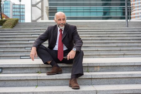 Cheerful grey haired business leader with smartphone enjoying work break outside. Elderly man in formal suit and tie sitting on outdoor office stairs. Mature businessman outside conceptの写真素材