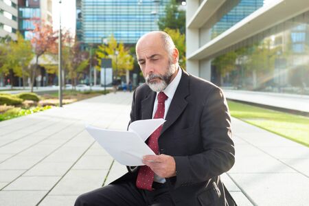 Serious mature businessman holding papers. Confident bearded businessman in formal wear sitting and holding documents on street. Paperwork conceptの写真素材