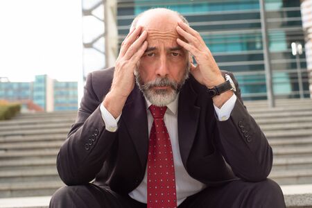 Ruined businessman sitting on steps and looking at camera. Front view of stressed mature businessman sitting with hands on head and looking at camera on street. Business problems conceptの写真素材
