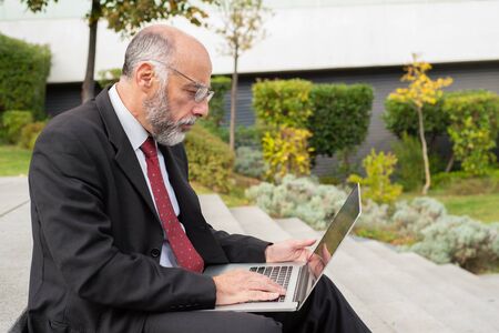 Serious businessman in eyeglasses using computer on street. Side view of focused mature businessman in eyeglasses sitting on steps and using laptop. Business and technology conceptの写真素材