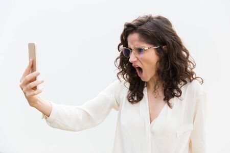Furious exaggerated woman in glasses staring at phone screen with open mouth. Wavy haired young woman in casual shirt standing isolated over white background. Communication conceptの写真素材
