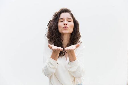 Cute positive student girl blowing air kiss. Wavy haired young woman in casual shirt standing isolated over white background. Sending kiss or romance conceptの写真素材