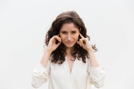 Stressed annoyed woman with covering ears with hands. Wavy haired young woman in casual shirt standing isolated over white background. Noise conceptの写真素材