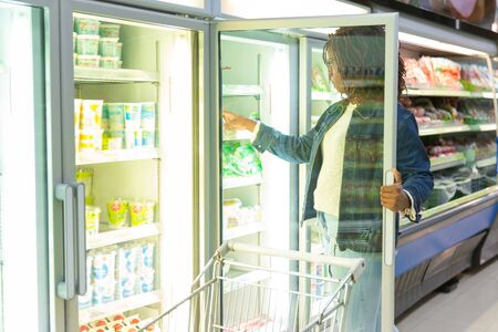 African American female customer taking package from fridge shelf in grocery store, choosing fresh food. Buyer shopping in supermarket. Grocery shopping conceptの写真素材