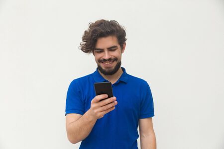 Happy positive guy using cellphone, looking at screen, laughing. Handsome bearded young man in blue...
