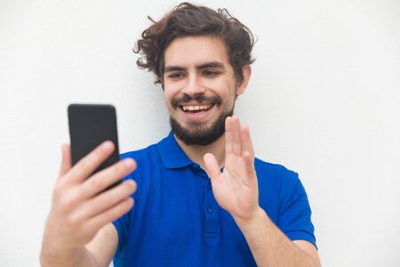 Joyful friendly guy with smartphone waving hello at screen. Handsome bearded young man in blue casual t-shirt posing isolated over white background. Video call conceptの写真素材