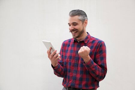 Excited man using digital tablet. Handsome hairy bearded man using tablet pc and triumphing on grey background. Success conceptの写真素材