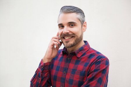 Joyful satisfied man talking on cell, looking at camera, smiling. Grey haired young man in checkered...