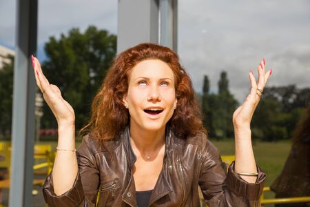 Smiling sarcastic woman sitting in outdoor coffee shop, rolling eyes, gesturing. Red curly haired...