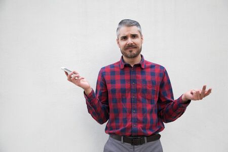 Doubtful man holding smartphone and looking at camera. Front view of uncertain bearded man in...
