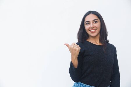 Cheerful positive woman pointing thumb away at copy space. Beautiful young woman in casual sweater posing isolated over white background. Presentation conceptの写真素材