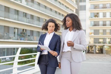 Serious female professional with documents consulting colleague on their way to office. Business women walking outside in city. Expertise conceptの写真素材