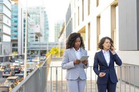 Business colleagues talking on cell and using tablet near office building. Business women working outside in city. Communication conceptの写真素材