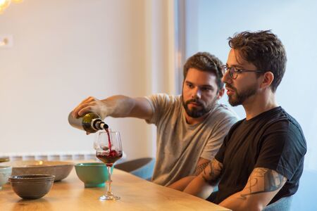 Handsome bearded man pouring wine in glass. Friends talking while sitting at table with wine and...