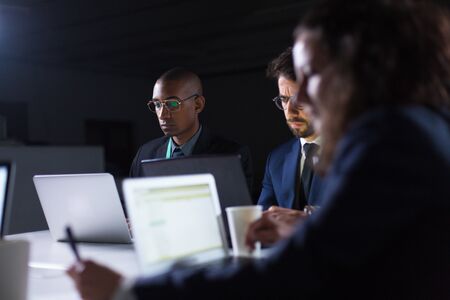 Confident people working over new project in dark office. Thoughtful business team sitting at long...