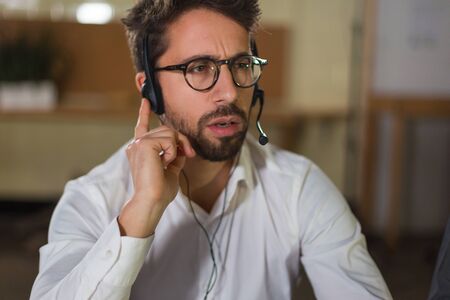 Thoughtful young man receiving incoming call. Front view of call center operator at work. Call...
