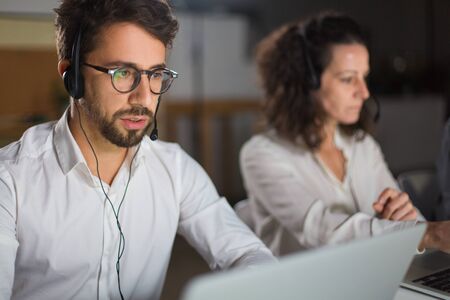 Front view of call center operator communicating with client. Confident young man in eyeglasses looking at screen of laptop and talking. Call center conceptの写真素材