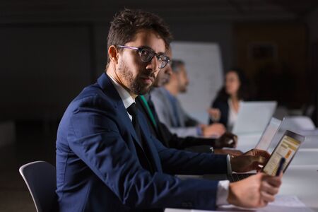Side view of focused man in eyeglasses working in office. Serious handsome businessman sitting at table with laptop and looking at camera. Business, working late conceptの写真素材