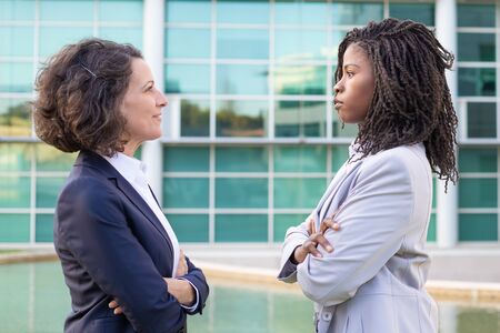 Coworkers standing with crossed arms and looking at each other. Side view of professional confident multiethnic businesswomen standing outside office building. Business conceptの写真素材