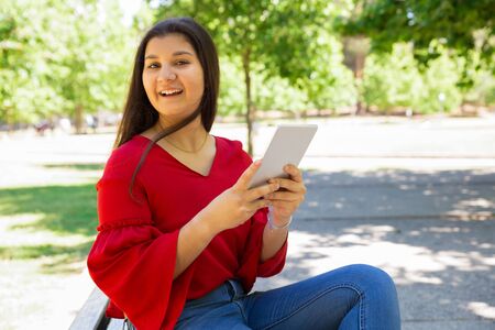 Smiling teen girl holding tablet while sitting on wooden bench. Cheerful teenager sitting in spring...